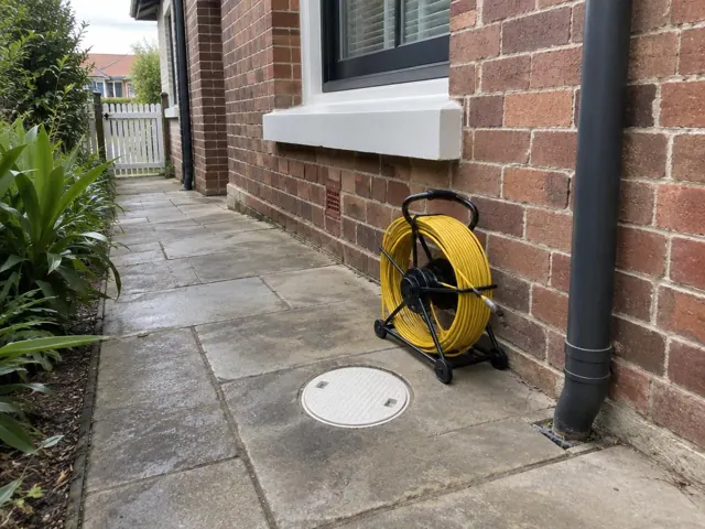 Capped drain inspection opening beside a coiled camera reel on a brick house side path in Clovelly