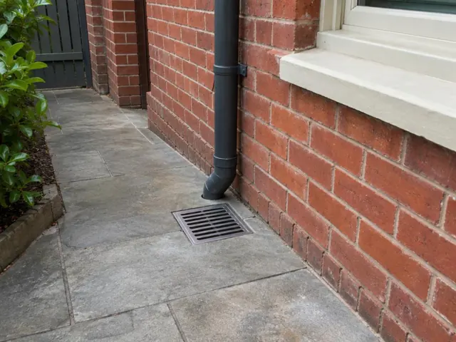 Outdoor gully grate set in paved walkway beside red brick wall and downpipe in Bondi