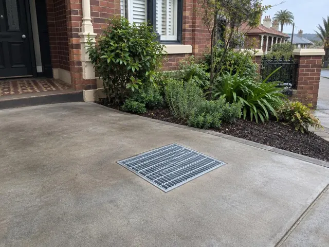 Concrete driveway with a flush stormwater grate beside brick house and garden in Bondi